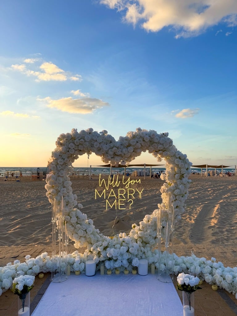 הצעת נישואין קשת לב פרחים לבנים עם שלט Will You Marry Me בחוף הים - Marryou